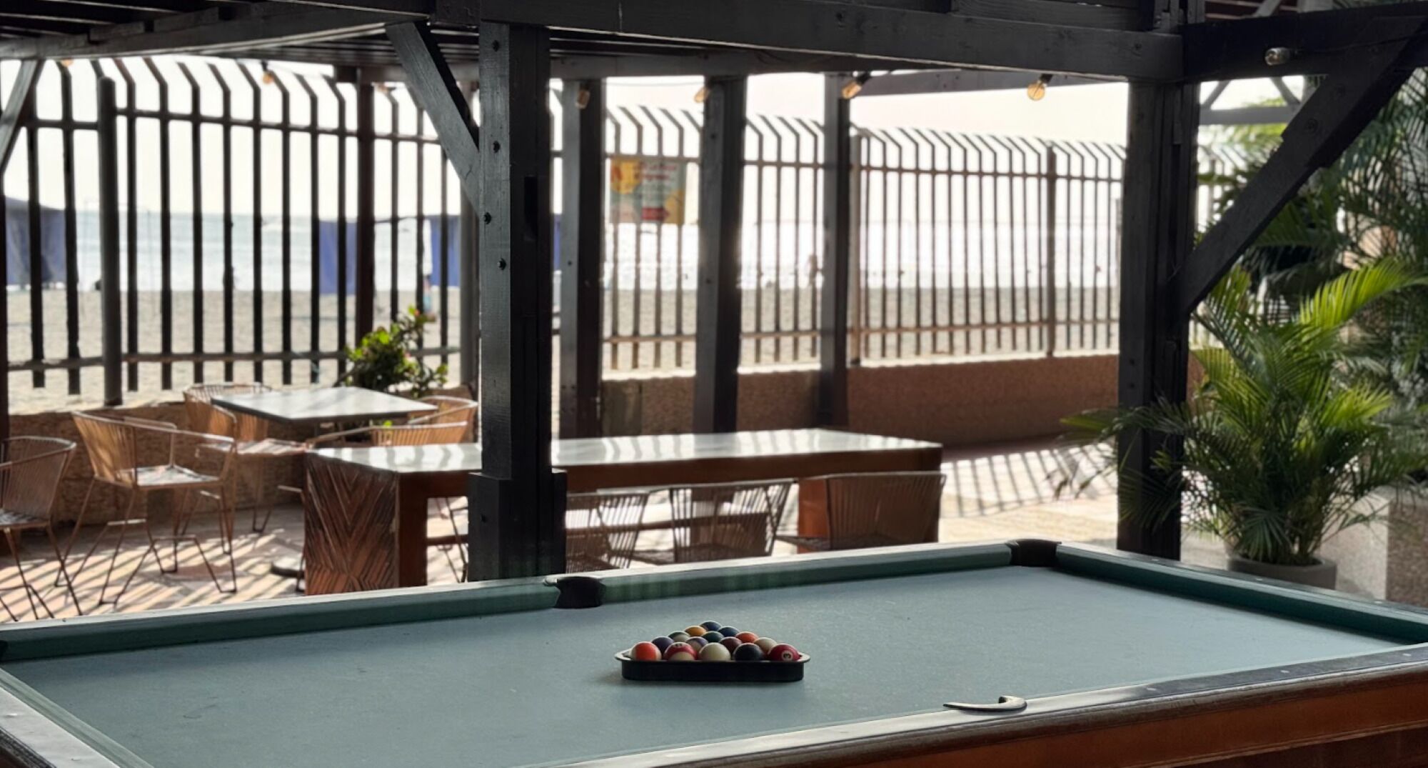 A pool table with balls in the center, surrounded by tables and chairs in an outdoor seating area with a metal fence and plants.