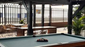 A pool table with balls in the center, surrounded by tables and chairs in an outdoor seating area with a metal fence and plants.