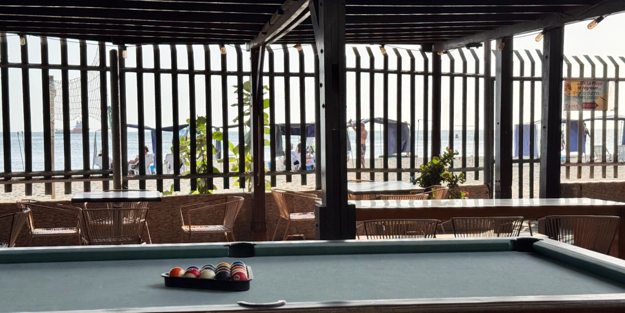 Una mesa de billar con un juego de bolas, rodeada de sillas y mesas de mimbre, con vista a la playa y al océano a través de una valla.