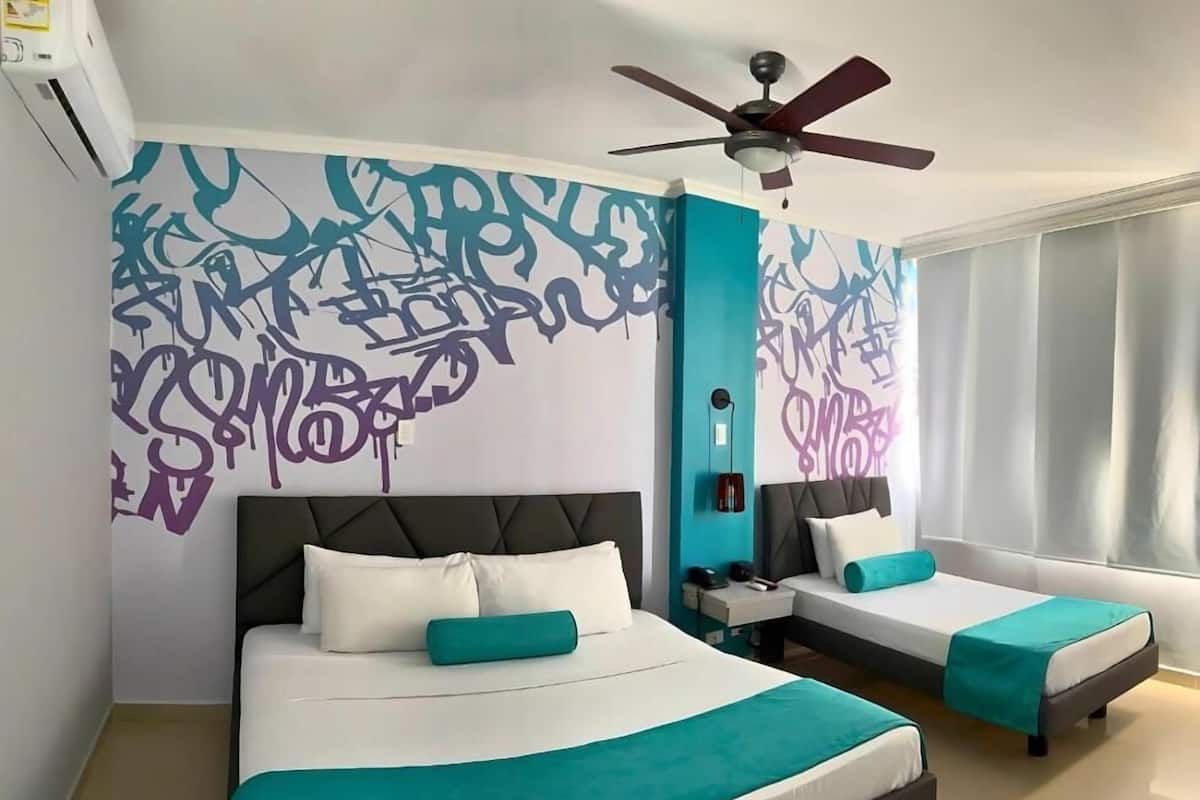 A hotel room with two beds, a ceiling fan, a window with curtains, and graffiti-style wall art.