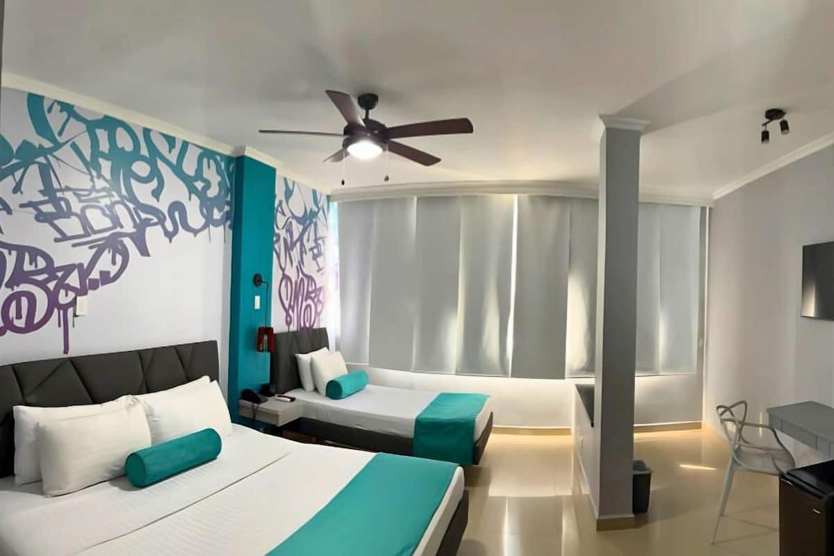 A modern hotel room with a large bed, a smaller bed, a desk, a TV, and a ceiling fan.