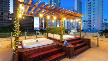 A rooftop terrace with wooden seating and hot tubs, surrounded by city buildings.