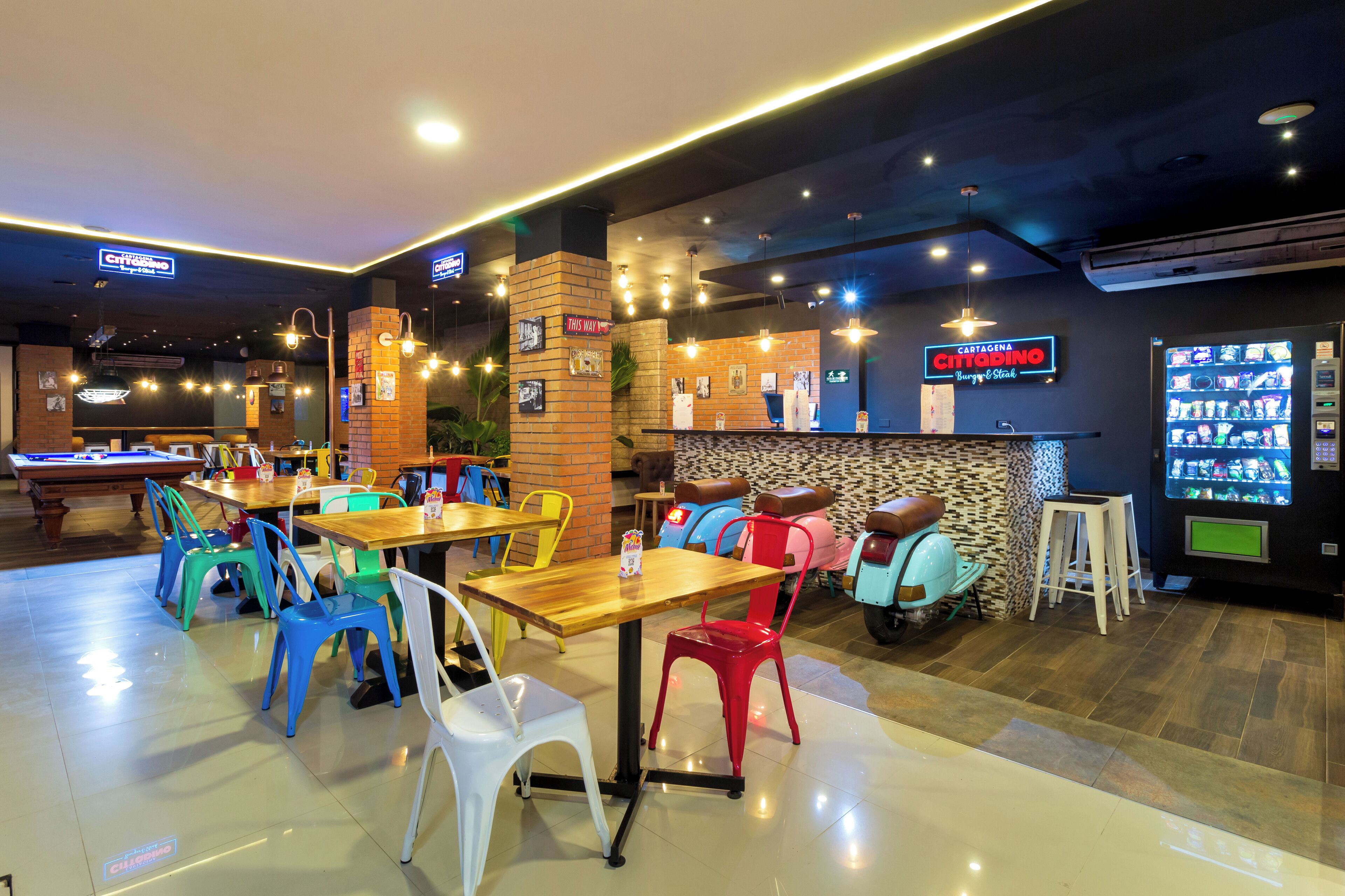 A restaurant with multiple tables and colorful chairs, a counter with a menu, and a vending machine.