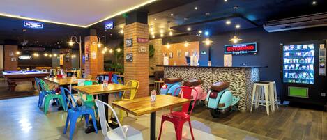 A restaurant with multiple tables and colorful chairs, a counter with a menu, and a vending machine.