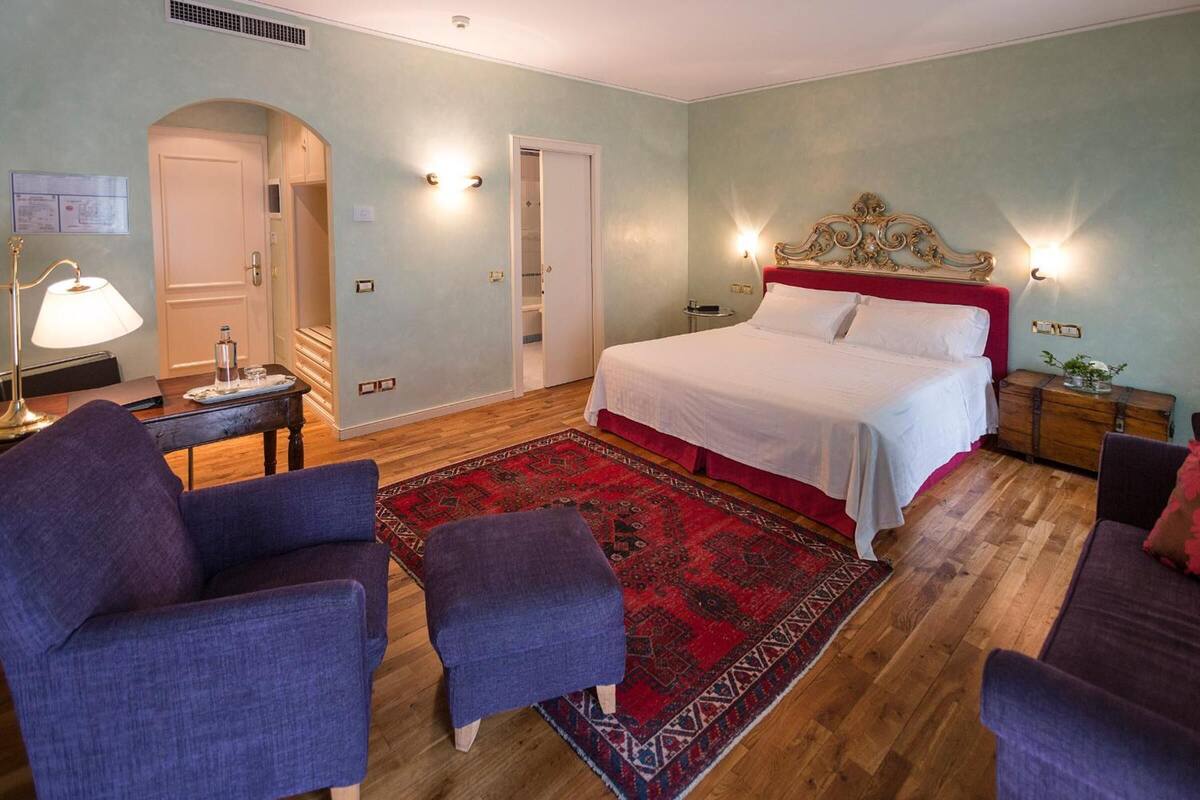 Ein Hotelzimmer mit einem Bett, zwei Sesseln, einem kleinen Tisch, einem Nachttisch, einem roten und schwarzen Teppich sowie einer weißen Tür.