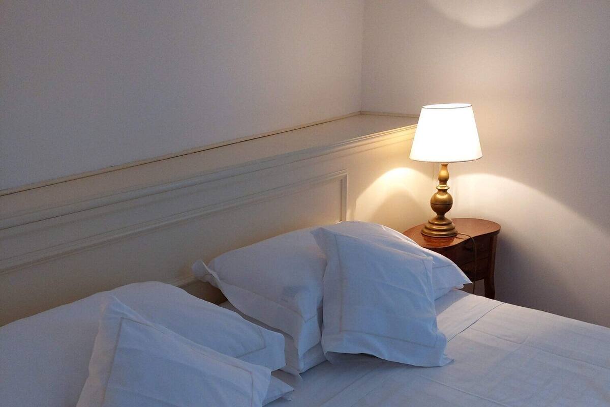 Ein ordentlich bezogenes Bett mit weißen Leinen und Kissen, ein Nachttisch mit einer Lampe sowie eine Wand mit schlichtem Design.