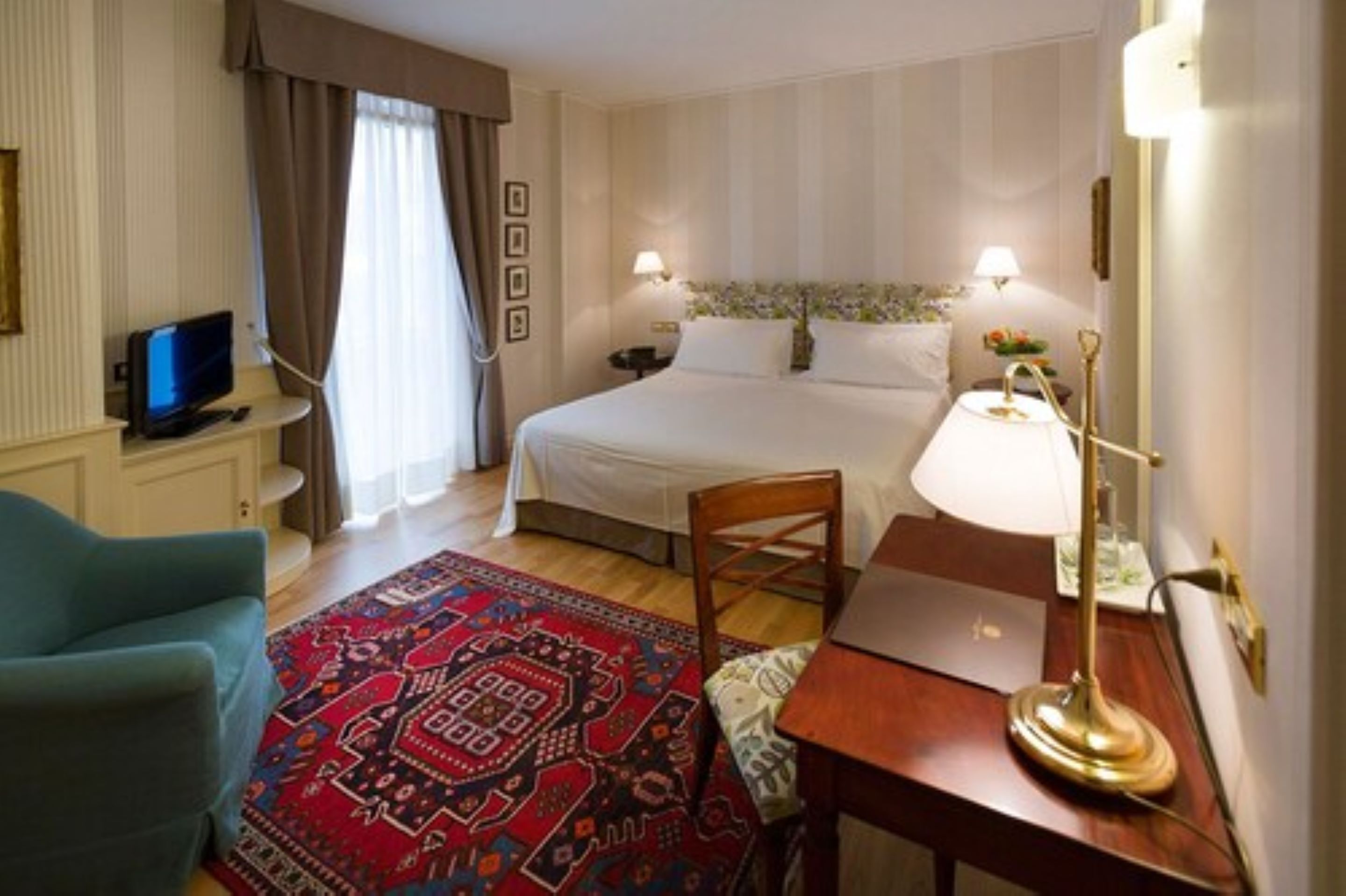A hotel room with a bed, bedside lamps, a desk, a chair, a TV, and a patterned rug.