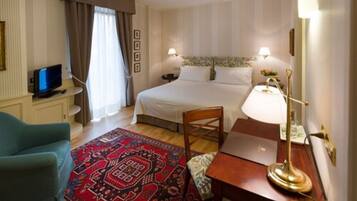 A hotel room with a bed, bedside lamps, a desk, a chair, a TV, and a patterned rug.