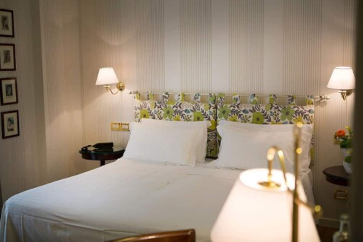 Ein Hotelzimmer mit einem großen Bett, zwei Nachttischlampen, einem blumigen Kopfteil und einem kleinen Tisch mit einem Telefon.