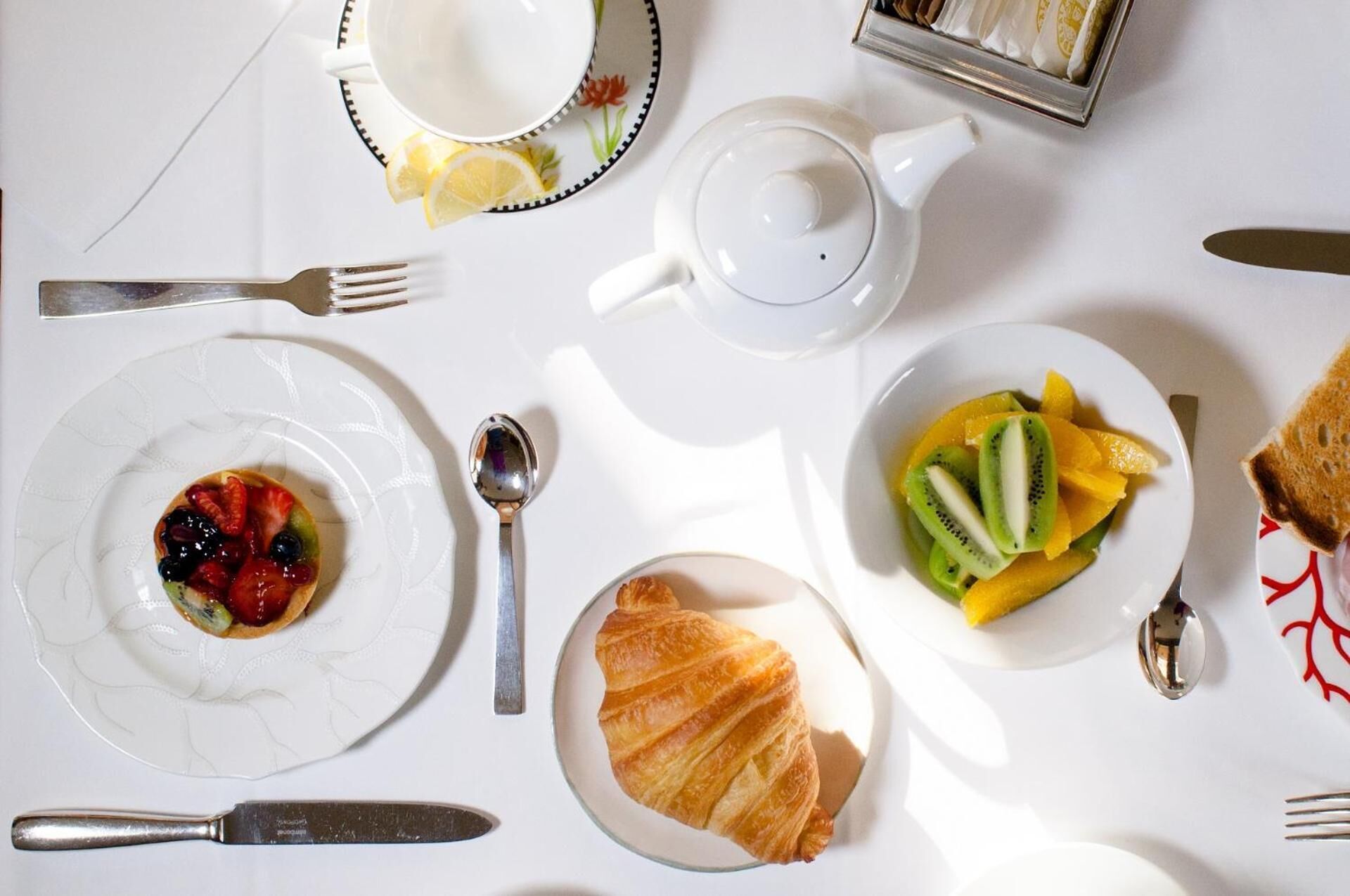 Disposição de café da manhã com um croissant, frutas, um bule e uma xícara em cima de uma mesa branca.