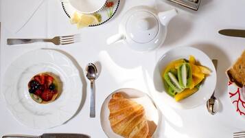 Disposição de café da manhã com um croissant, frutas, um bule e uma xícara em cima de uma mesa branca.
