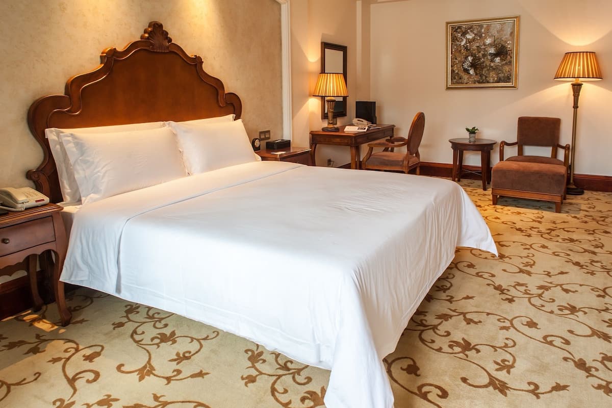 Habitación de hotel con una cama grande, una mesita de noche de madera con teléfono, un escritorio con lámpara, una silla y una pequeña mesa con una planta.