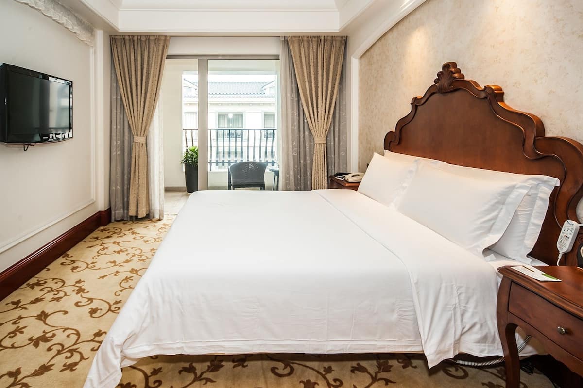 Habitación de hotel con una cama grande, una mesita de noche con lámpara, un televisor montado en la pared y vistas al balcón exterior.