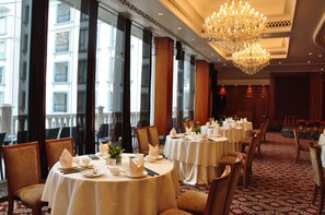 Ein Speisesaal eines Hotels mit runden Tischen, großen Fenstern und einer Chandelüster.
