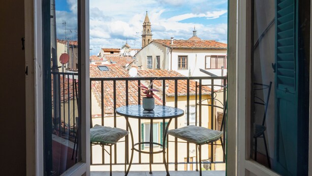 Een open balkon met een tafel en stoelen, uitzicht op gebouwen met rode dakpannen en een heldere lucht.