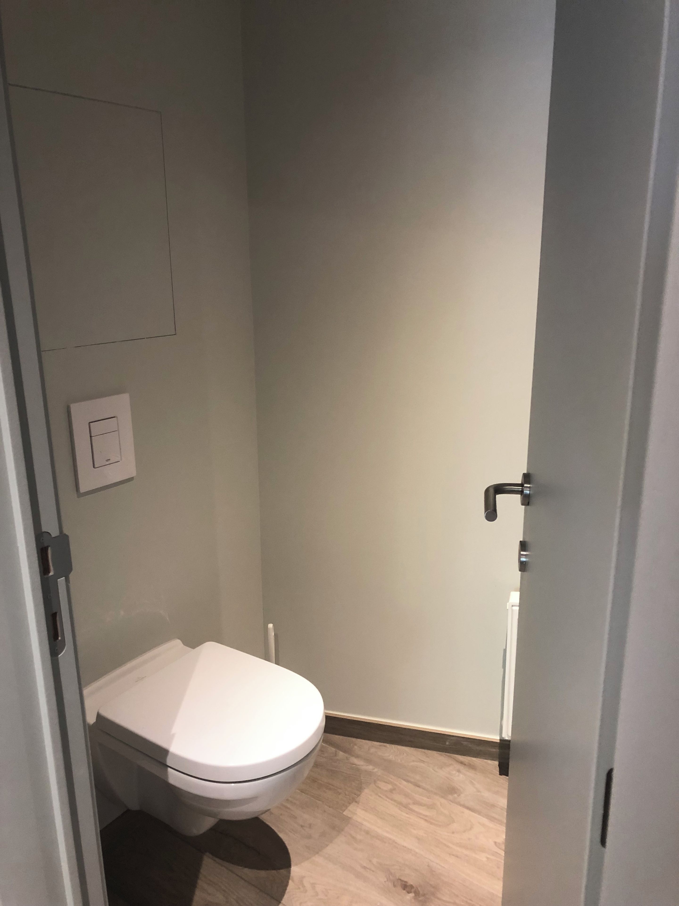 Une petite salle de bain fermée, équipée de toilettes, d’un interrupteur fixé au mur et d’une porte.