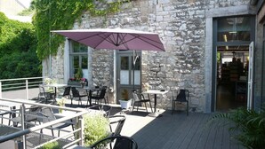 Une terrasse extérieure avec des tables et des chaises, un parasol violet et un bâtiment en pierre doté d’une porte en verre.