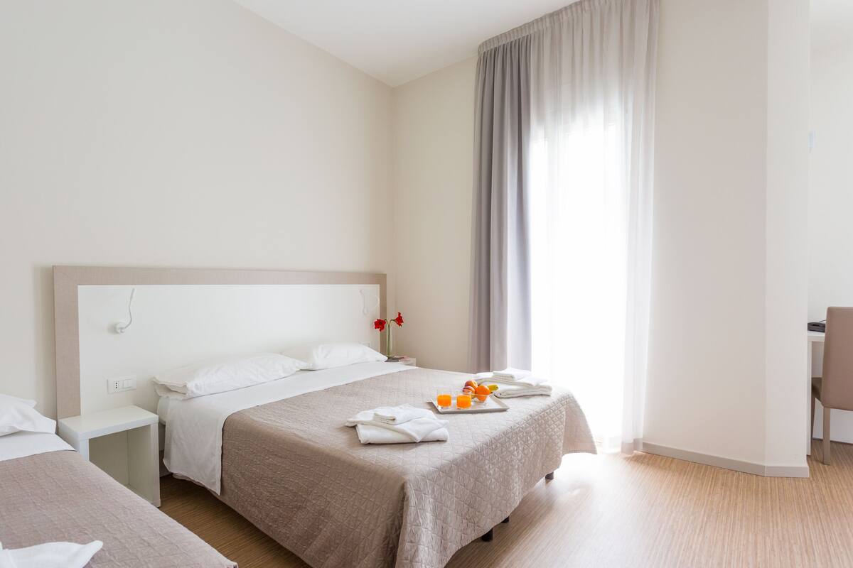 Quarto de hotel com duas camas, uma mesa de cabeceira com abajur, uma janela com cortinas e uma bandeja com toalhas e sumo de laranja.