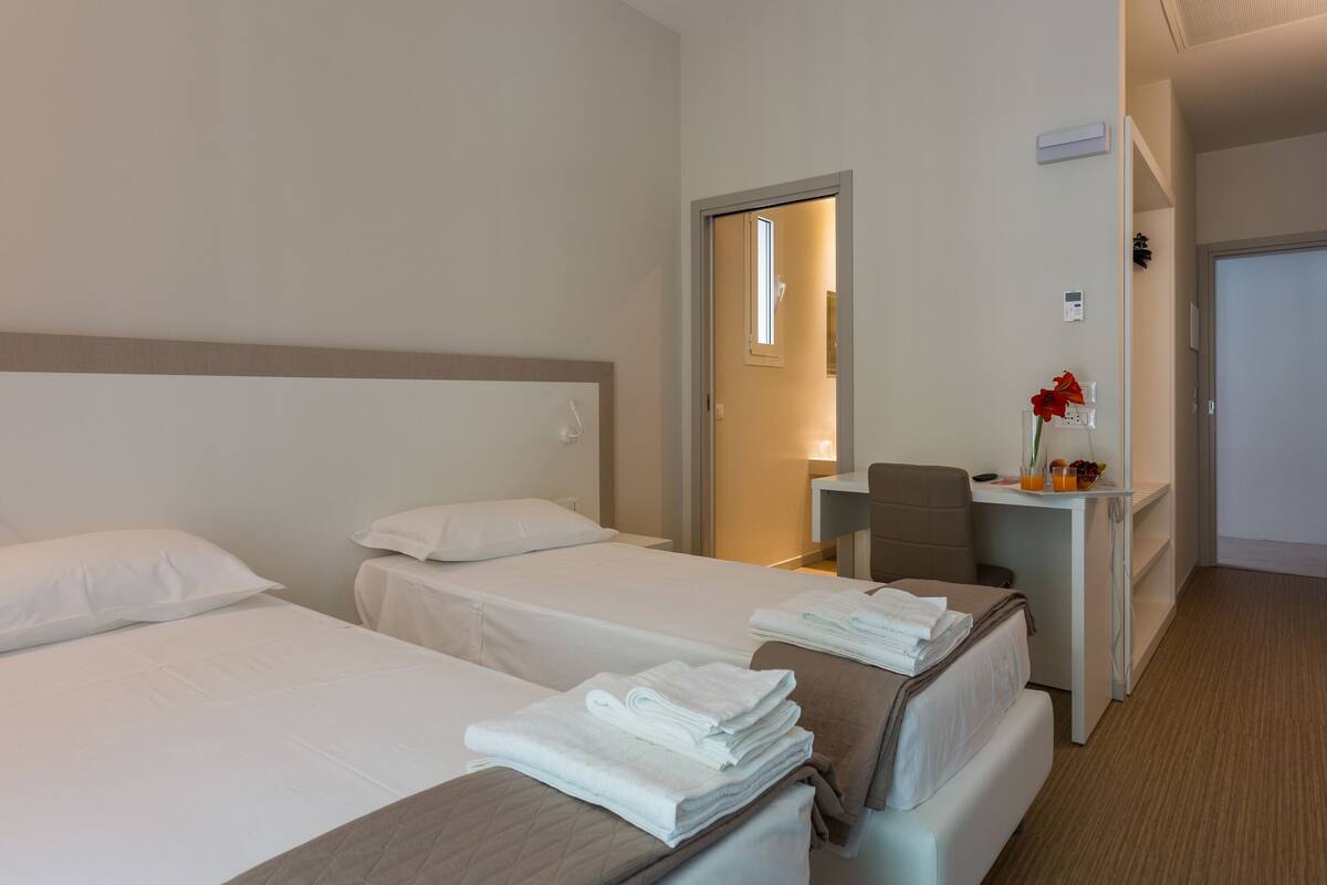 Quarto de hotel com duas camas, uma escrivaninha com cadeira e banheiro visível através de uma porta aberta.