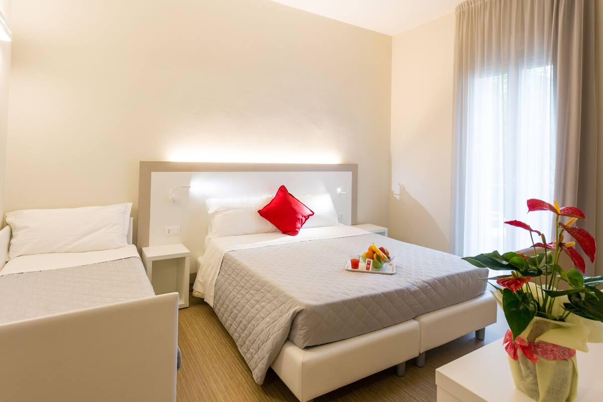 Quarto de hotel com duas camas de solteiro, uma cama grande, uma mesa de cabeceira com um travesseiro vermelho, uma mesinha com uma cesta de frutas e uma chávena, e um vaso com flores.