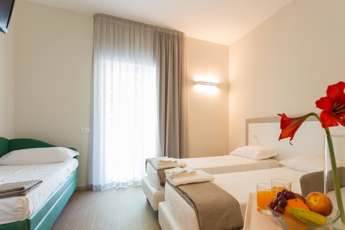 Quarto de hotel com duas camas, uma janela grande com cortinas leves, mesa de cabeceira com um vaso de flores e uma bandeja de frutas.