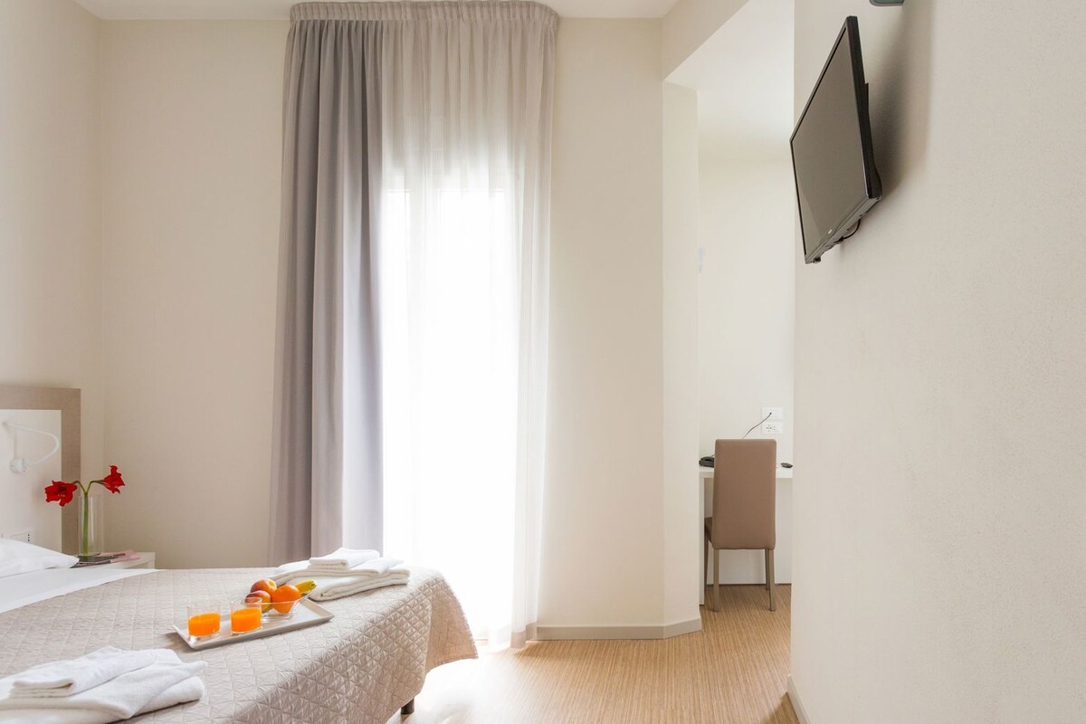 A hotel room with a bed, a desk, a chair, a TV mounted on the wall, and a window with curtains.