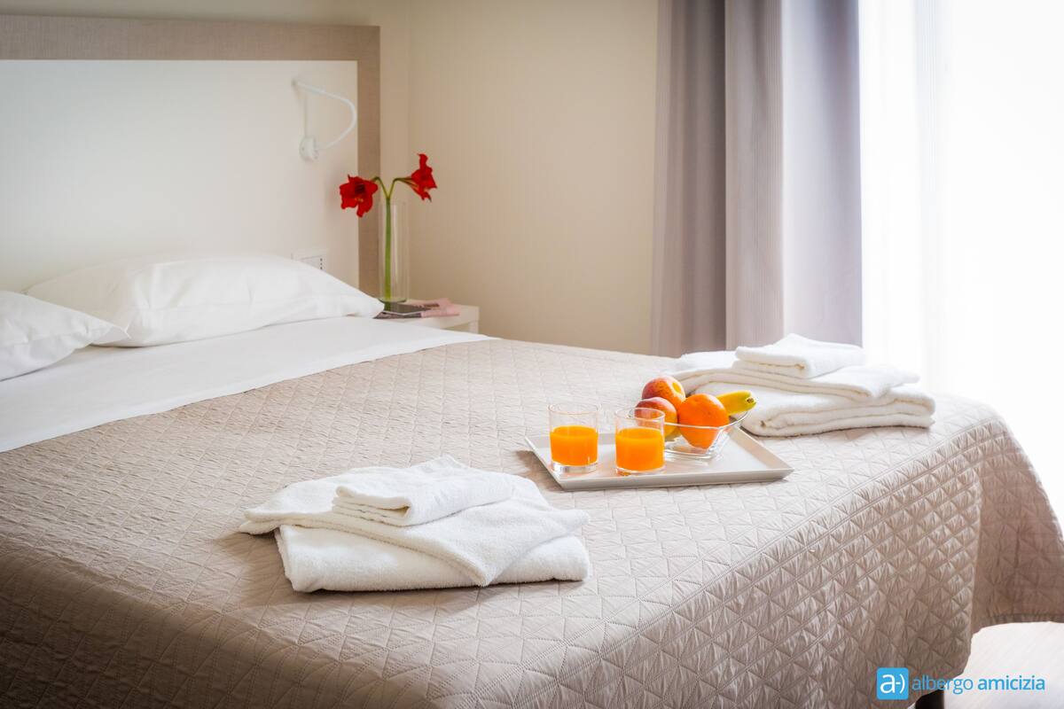 Quarto de hotel com uma cama, uma bandeja com suco de laranja e frutas, e um vaso com flores vermelhas na mesa de cabeceira.