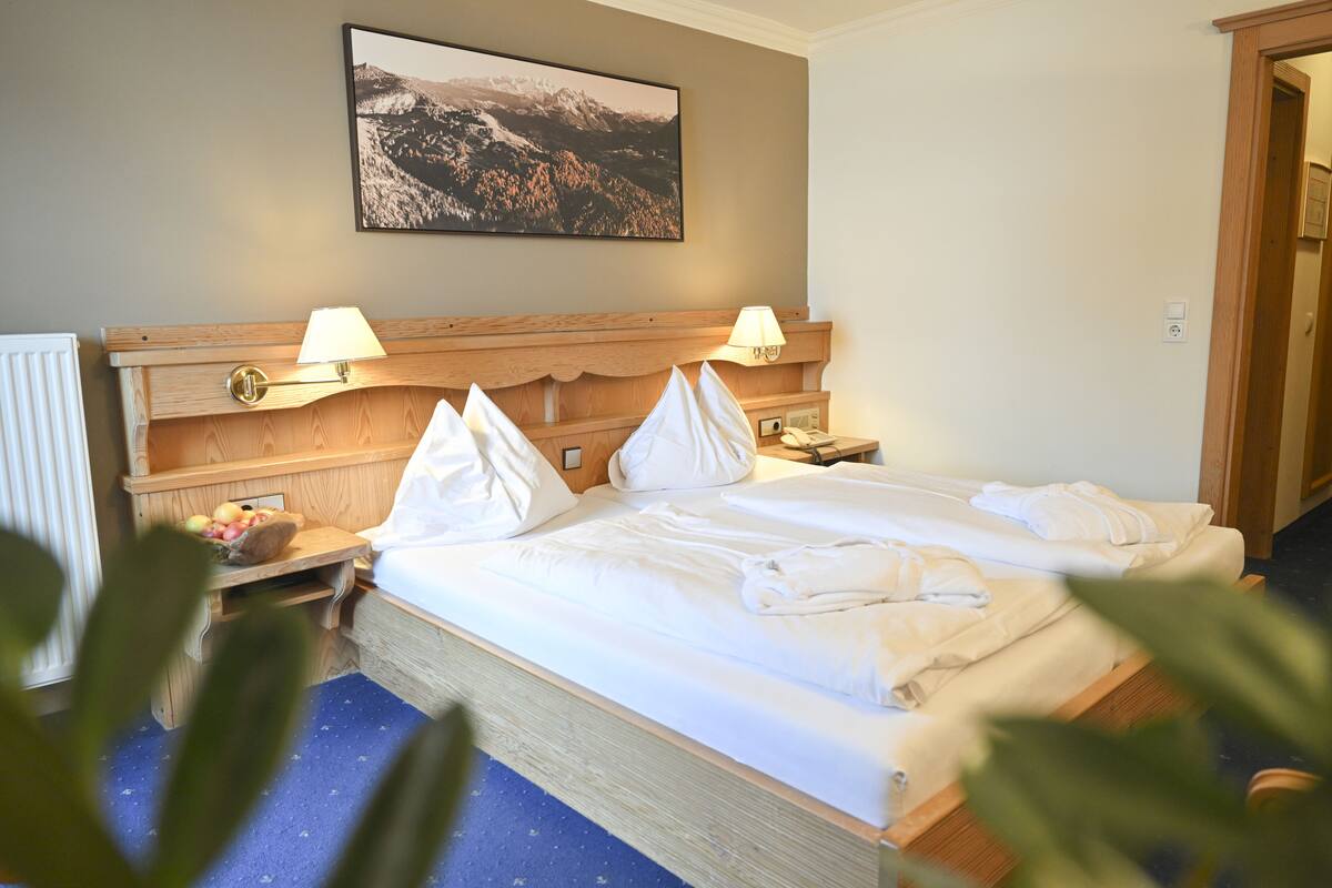 A hotel room with a wooden headboard, two bedside lamps, a neatly made bed with pillows and a blanket, a fruit bowl on the nightstand, and a framed picture on the wall.