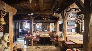 Un interior rústico con vigas de madera, piso de baldosas de piedra y una zona central de estar con bancos y mesas.