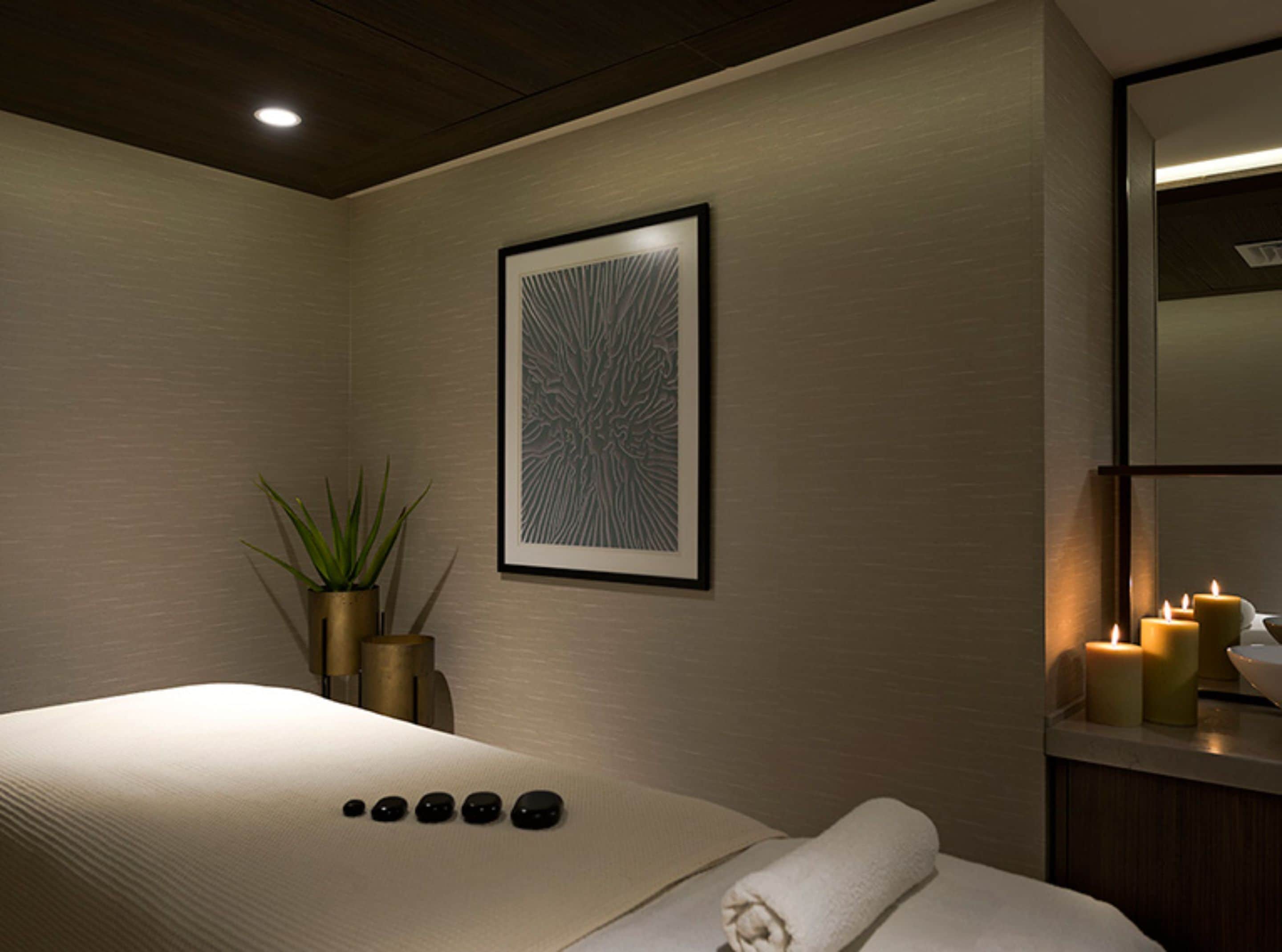 A modern spa room with a bed, a wall-mounted artwork, a potted plant, and candles.