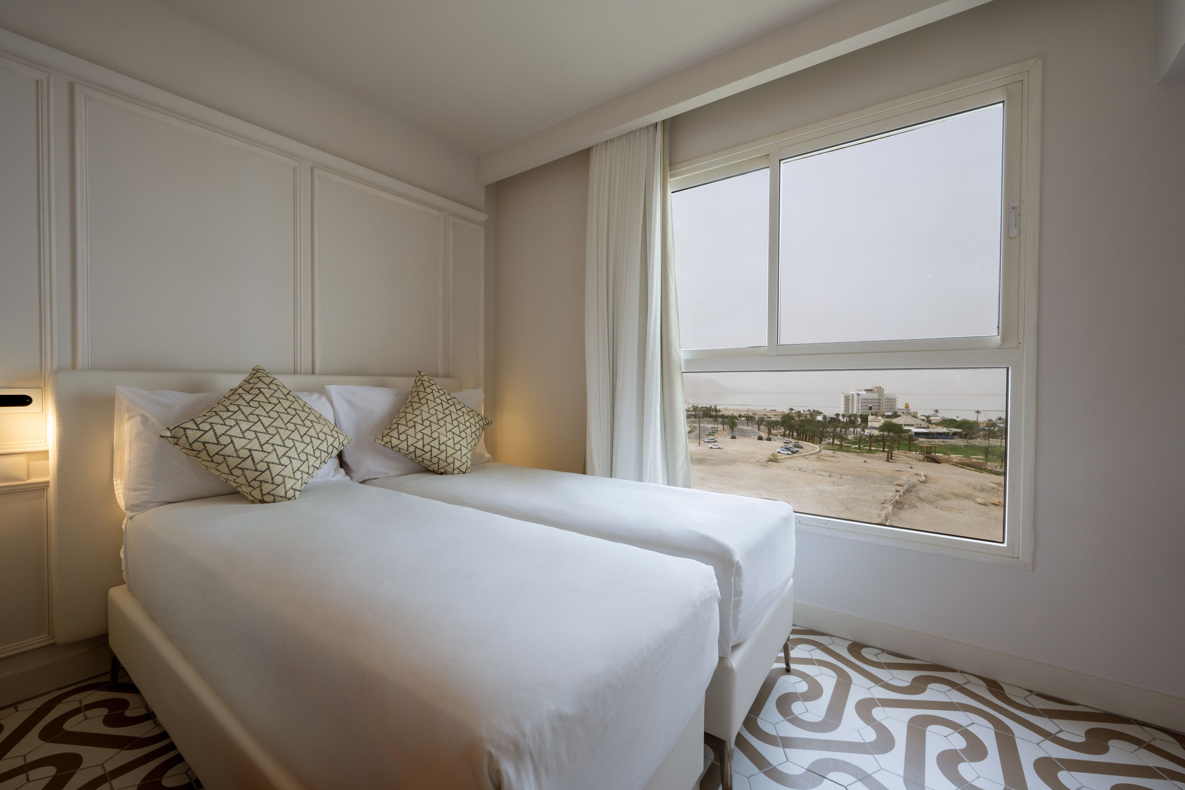 Habitación de hotel con dos camas, un ventanal con vistas a la playa y edificios, y un suelo estampado.