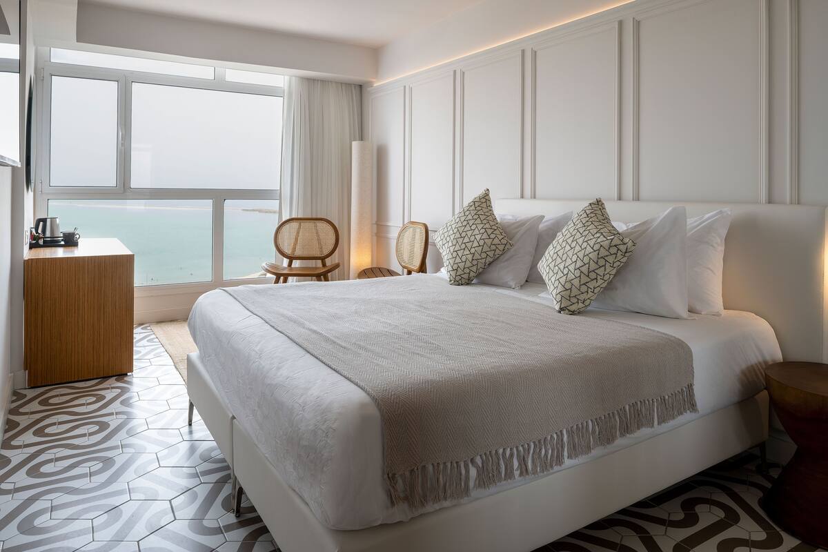 A hotel room with a large bed, a wooden desk, and a view of the sea through a large window.