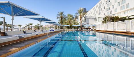 A swimming pool with lounge chairs and umbrellas, surrounded by palm trees and a hotel building.