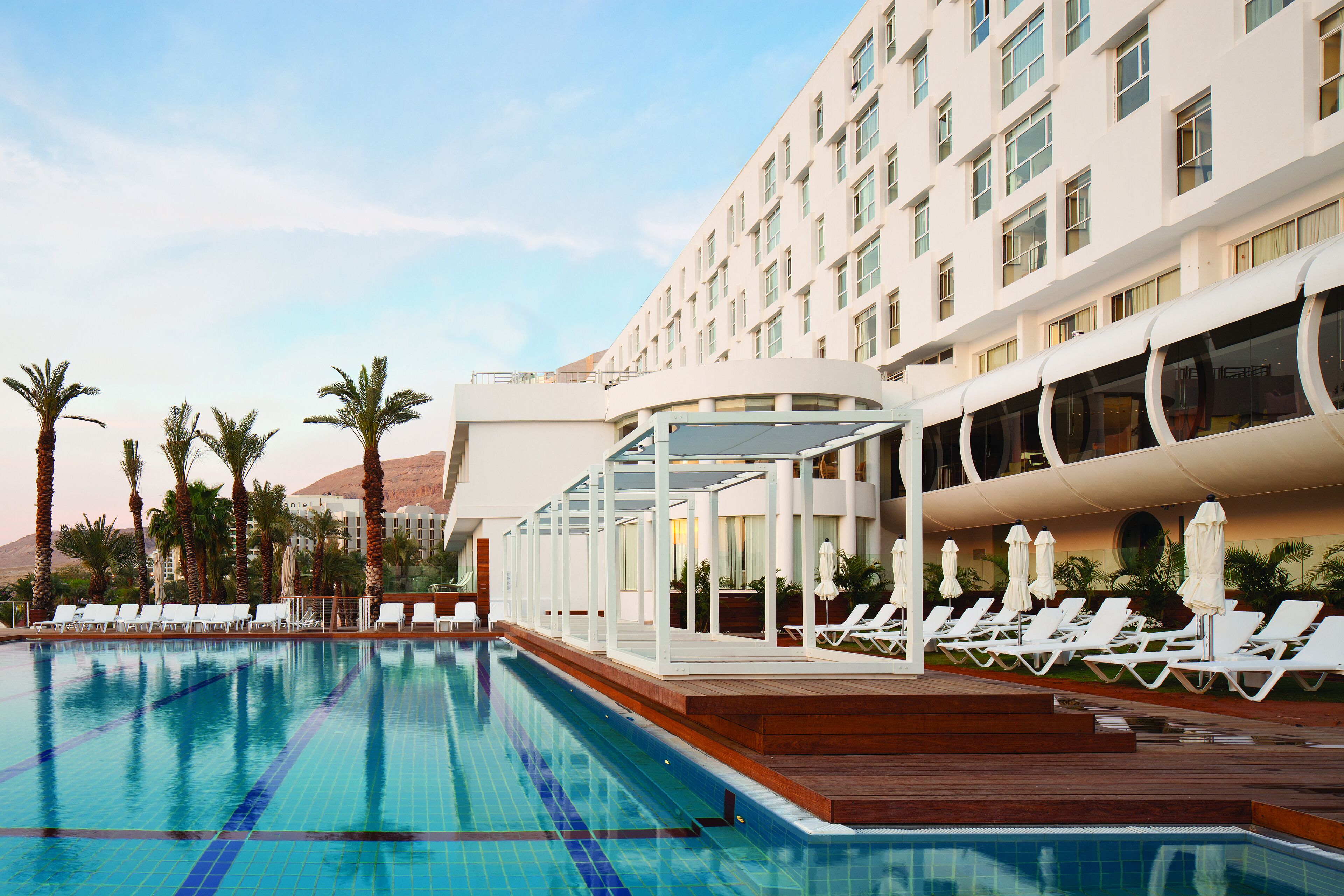 A swimming pool with lounge chairs and umbrellas, surrounded by palm trees and a hotel building.