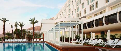A swimming pool with lounge chairs and umbrellas, surrounded by palm trees and a hotel building.