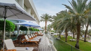 A sun deck with lounge chairs and umbrellas, surrounded by palm trees and a glass railing. - Noga By Isrotel Collection (Tamar)