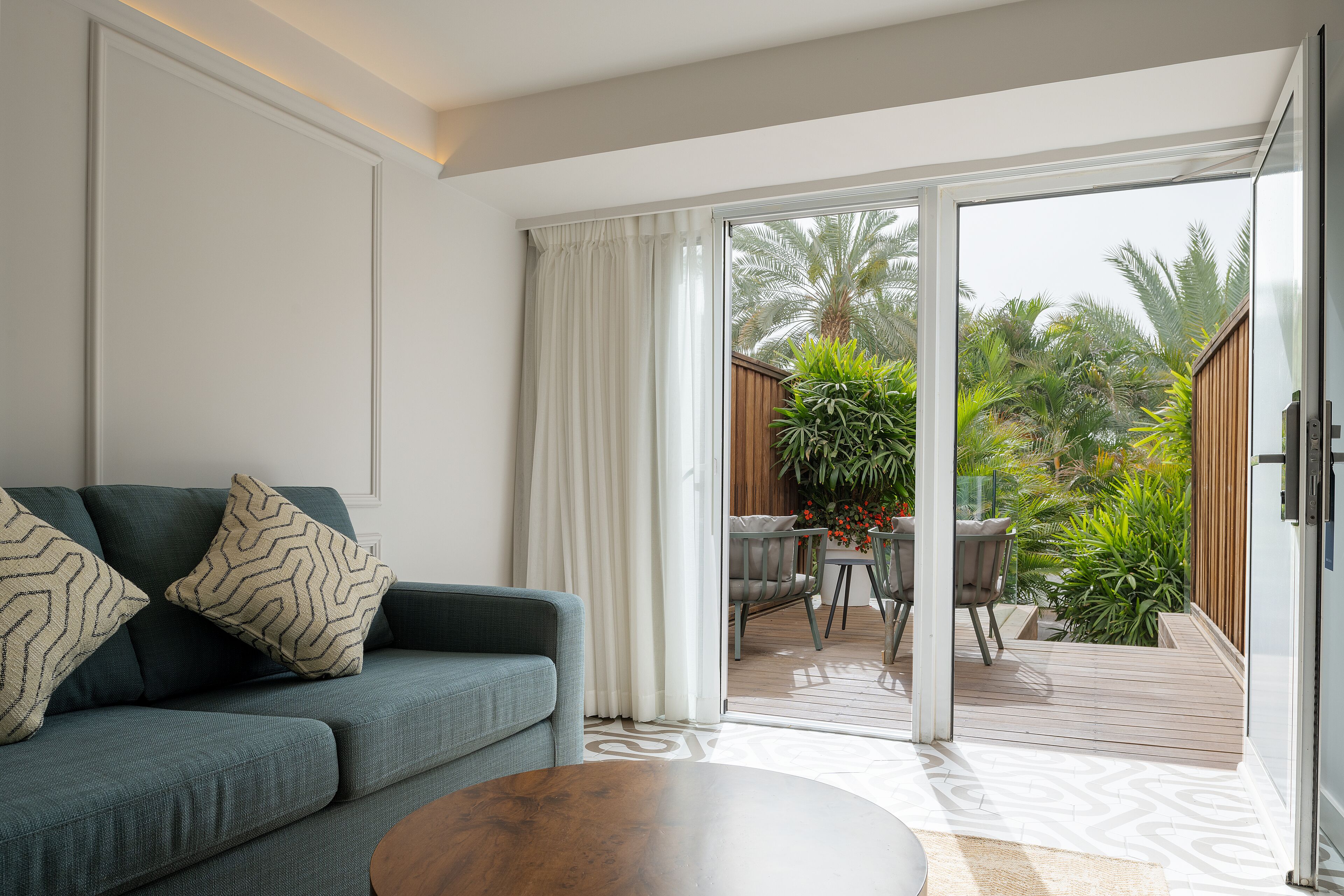 Una sala de estar moderna con un sofá, una mesa de centro y un ventanal corredero que da a un patio exterior con terraza de madera y plantas.