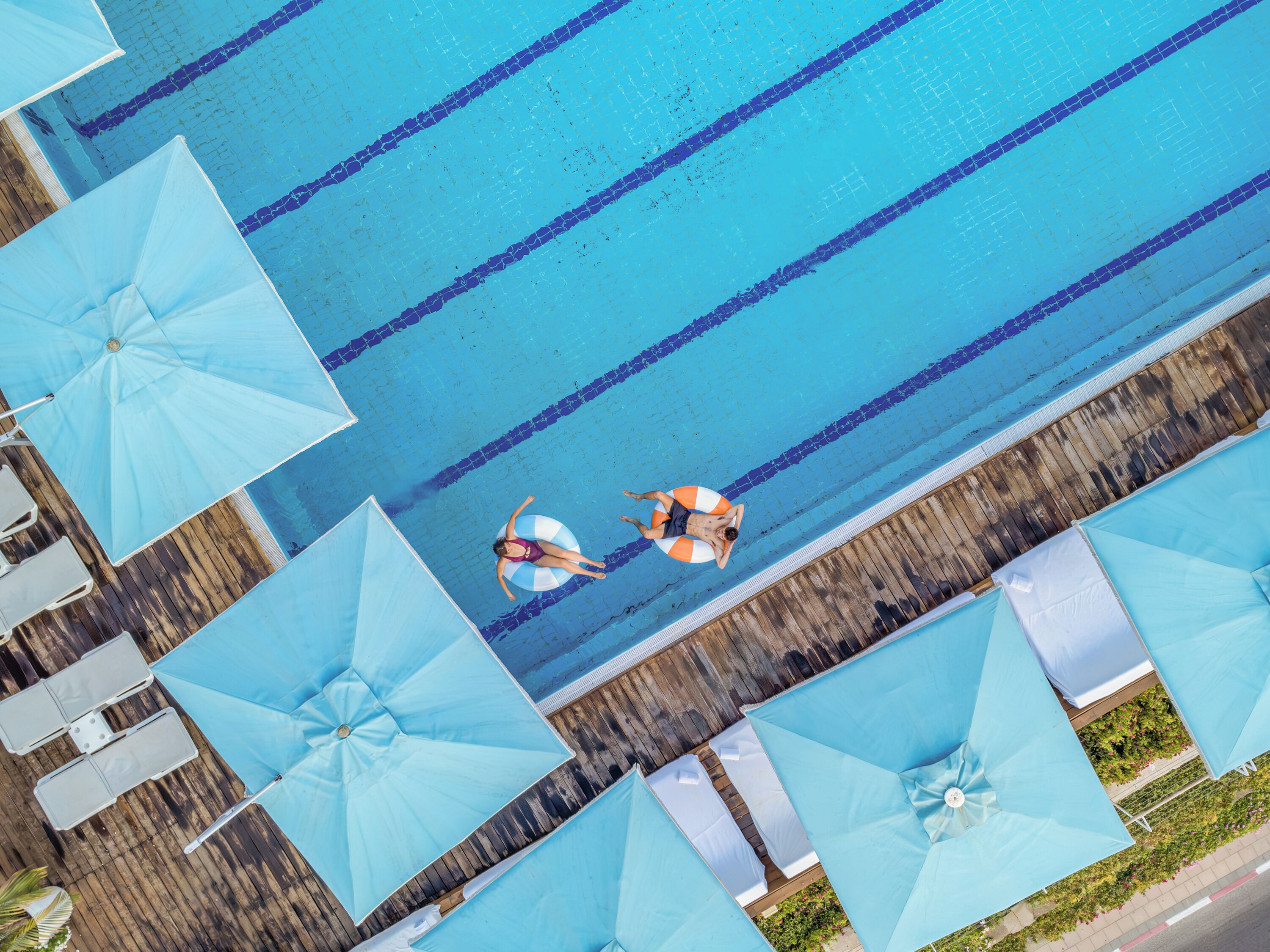 An aerial view of a swimming pool with multiple lanes, surrounded by blue umbrellas and lounge chairs.