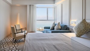A hotel room with a large bed, a blue sofa, a chair, and a window with a view of the city. - Noga By Isrotel Collection (Tamar)