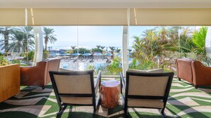 A room with two chairs facing a pool area, a wooden table, and a view of the pool and palm trees. - Noga By Isrotel Collection (Tamar)