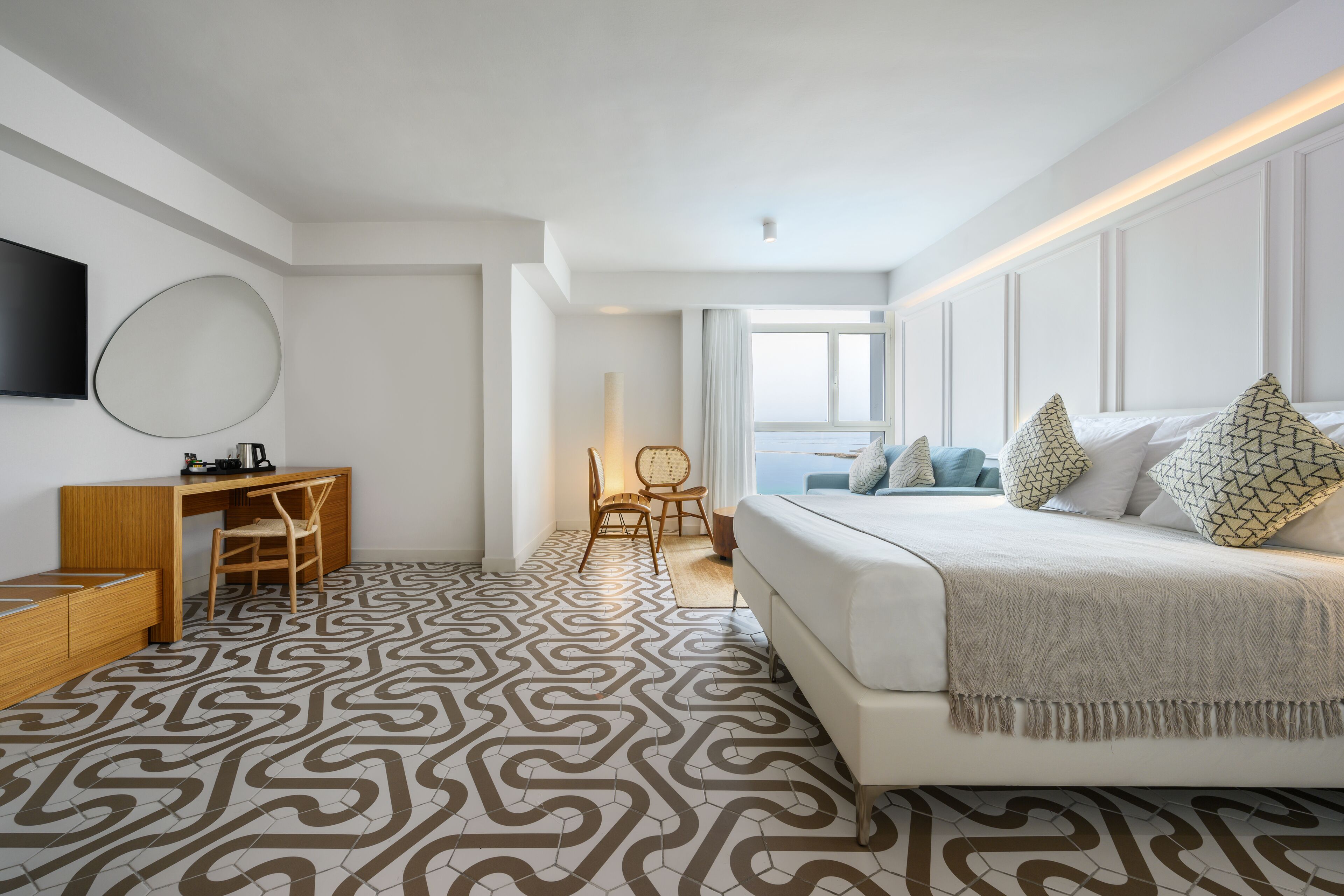 A modern hotel room with a large bed, a desk with a chair, a TV mounted on the wall, and a patterned floor.