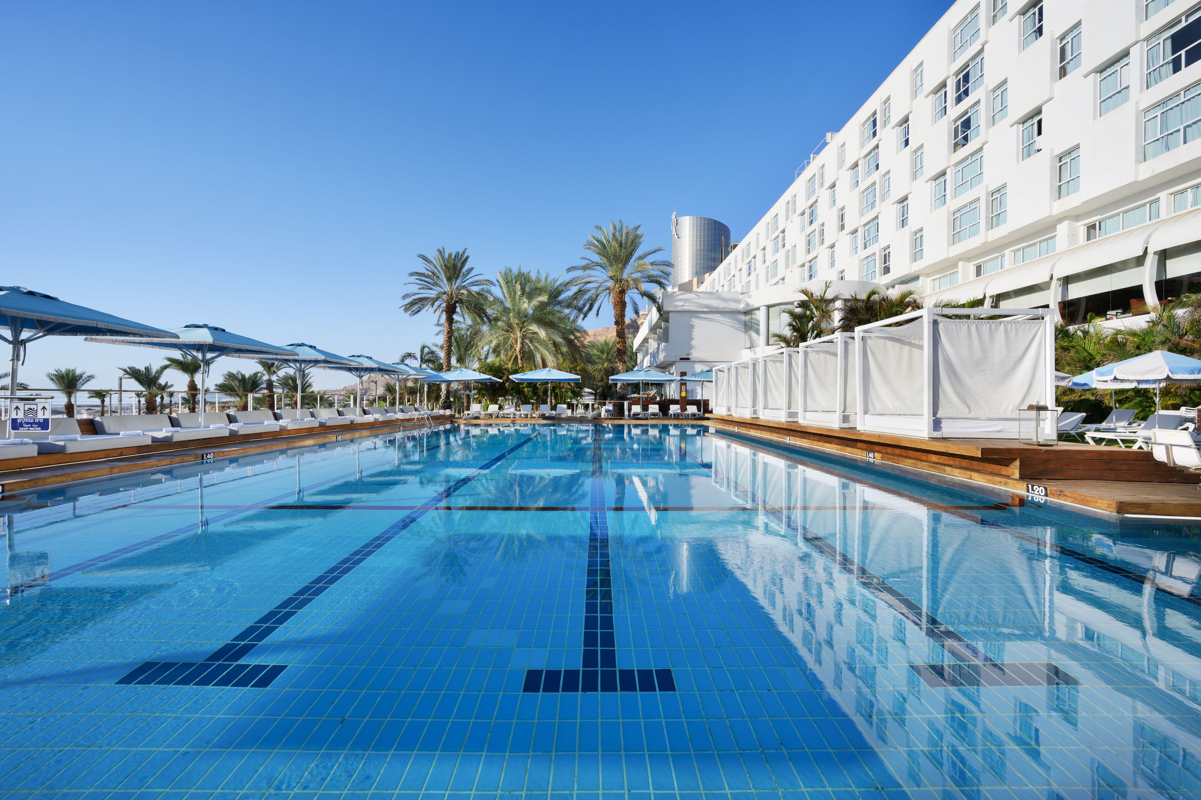 Una piscina con azulejos azules cristalinos, rodeada de sillas de descanso y sombrillas, con un edificio de hotel al fondo.