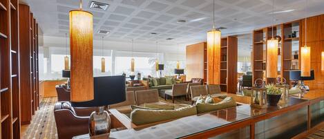 A spacious hotel lobby with a modern design, featuring a large seating area, wooden bookshelves, and a glass partition.