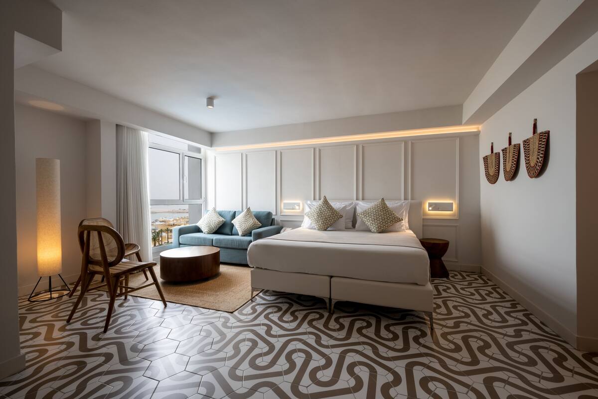 A modern bedroom with a large bed, a blue sofa, a wooden chair, and a patterned floor.