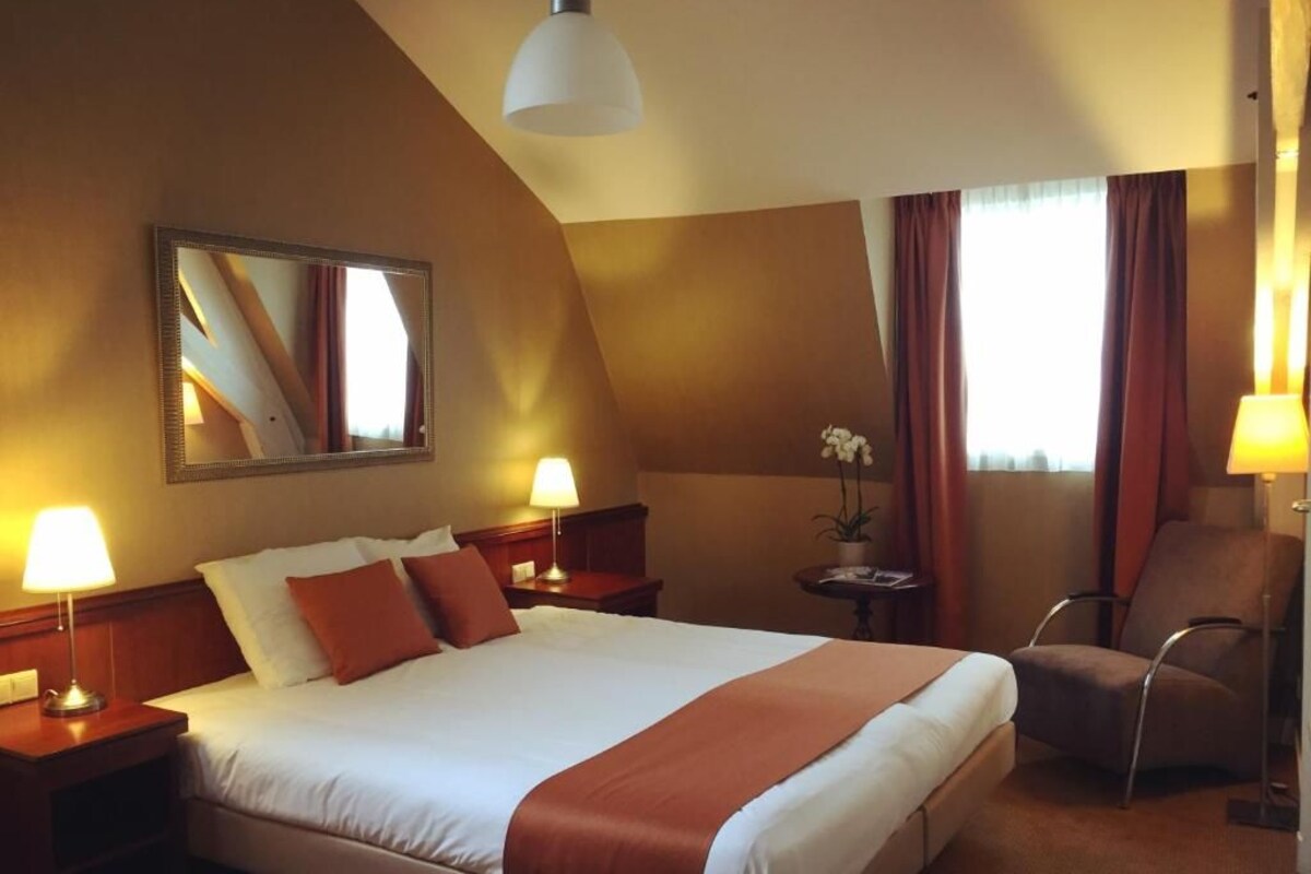 Een hotelkamer met een groot bed, twee bedlampjes, een stoel, een kleine tafel met een plant en een spiegel aan de muur.