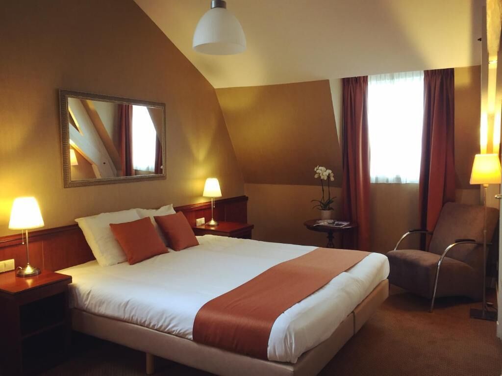 A hotel room with a large bed, two bedside lamps, a chair, a small table with a plant, and a mirror on the wall.