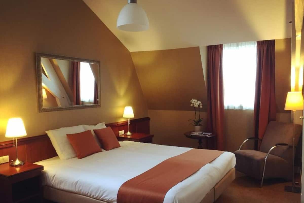 Habitación de hotel con una cama grande, dos lámparas de noche, una silla, una mesita con una planta y un espejo en la pared.