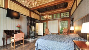 A spacious bedroom with a large bed, a wooden desk with a piano, a chandelier, and a television mounted on the wall.