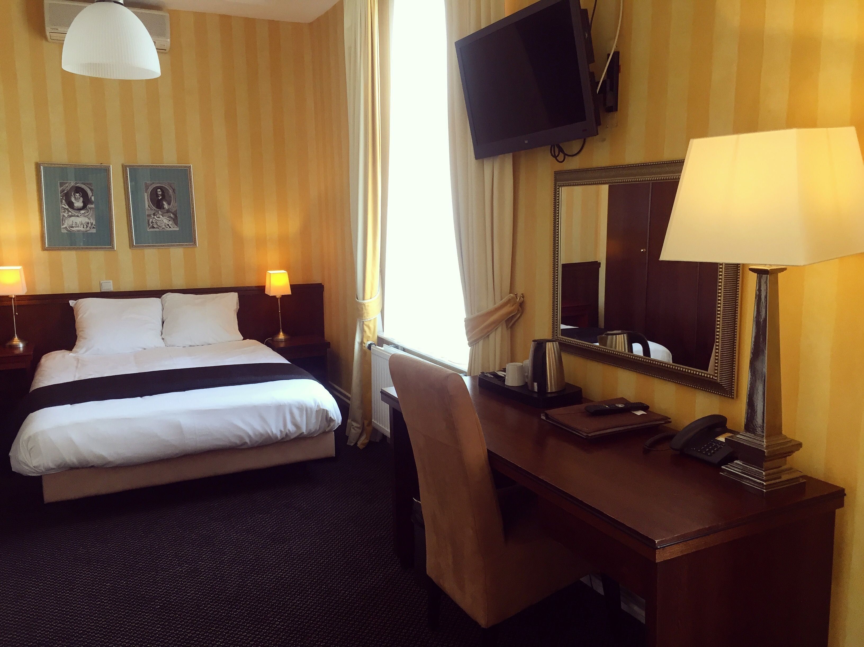 A hotel room with a bed, bedside tables, a desk with a chair, a television, and a mirror.