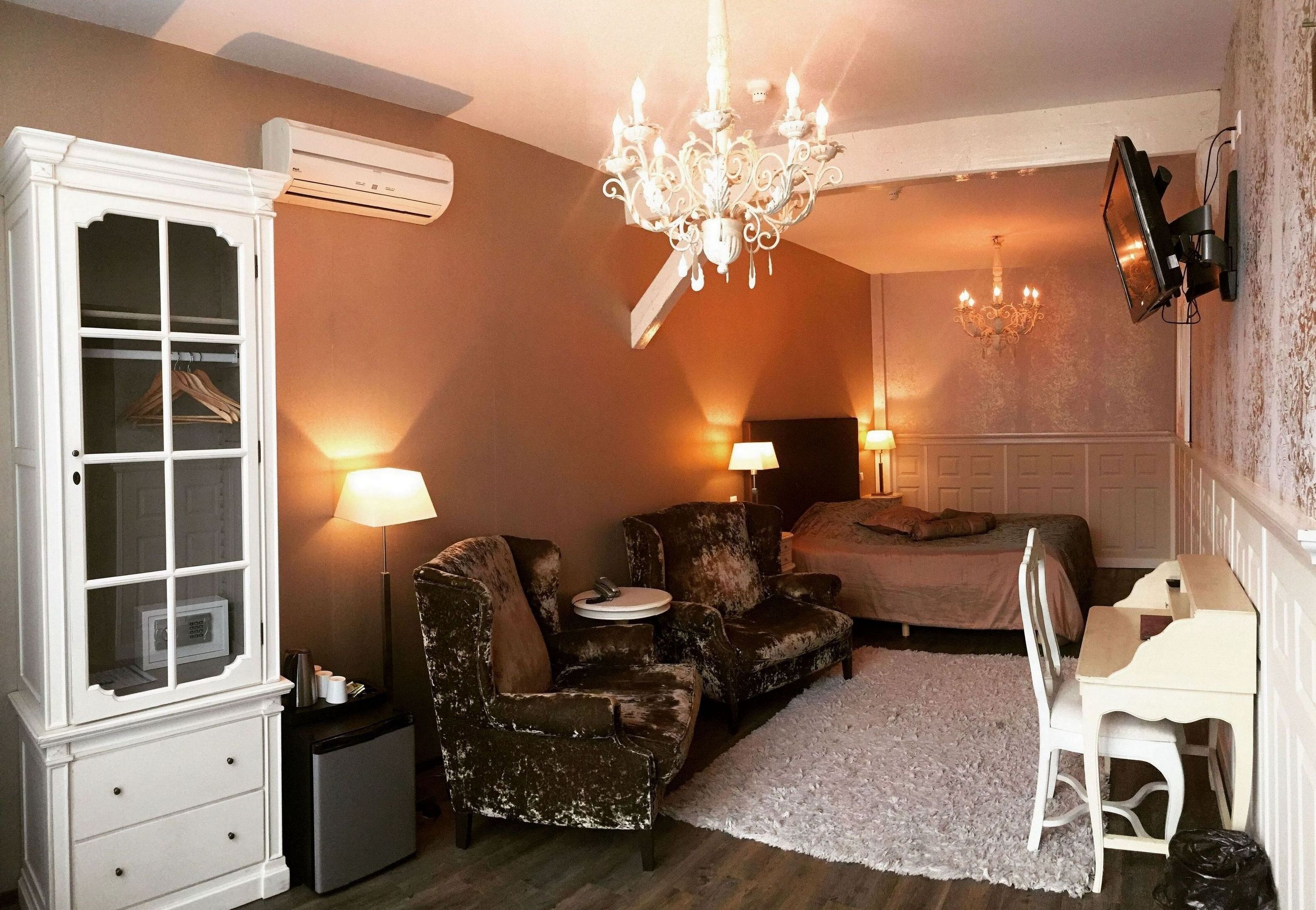 A bedroom with a bed, two chairs, a desk, a chandelier, and an air conditioning unit.